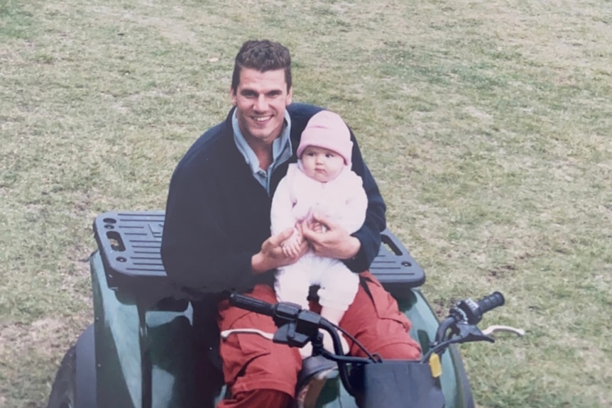 A young man holding a baby on a four-wheeled motorcycle.