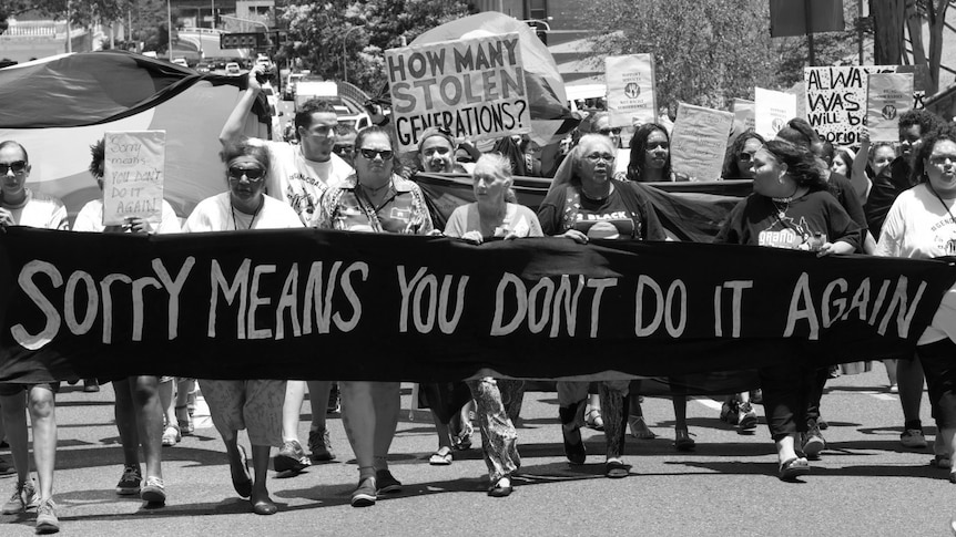 A group of older women walk the streets with a large banner that says 'Sorry means you don't do it again'