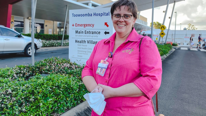 A woman stands in front of a hospital with a mask in hand.