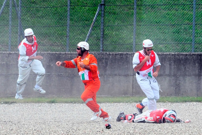 A man in an orange jumpsuit and helmet runs away from Michele Pirro lying prone on gravel, two other rush towards him