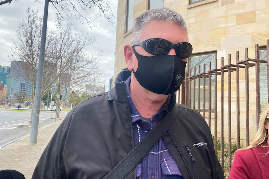 A man with grey wearing a face mask and sunglasses leaves court surrounded by journalists