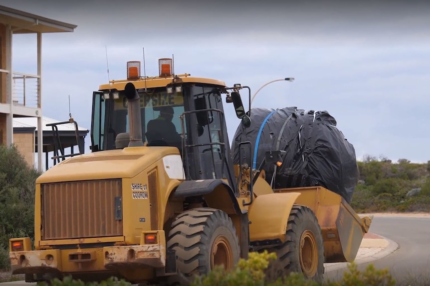 A large object wrapped in plastic being driven through residential streets on a front end loader