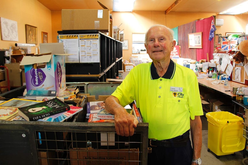 An elderly man in a high-vis shirt leans on a cage of books.