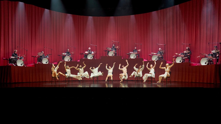 Nine dancers in white costumes strike abstract poses seated on a stage. Behind them, nine drummers in black play on a podium.
