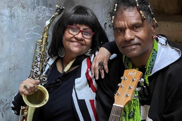 an Aboriginal woman with short, straight black hair holding a saxophone next to an Aboriginal man with dreadlocks and a guitar