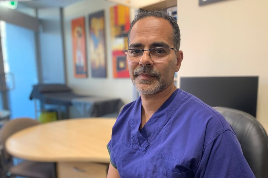 A man wearing glasses and hospital scrubs, smiles for the camera. 