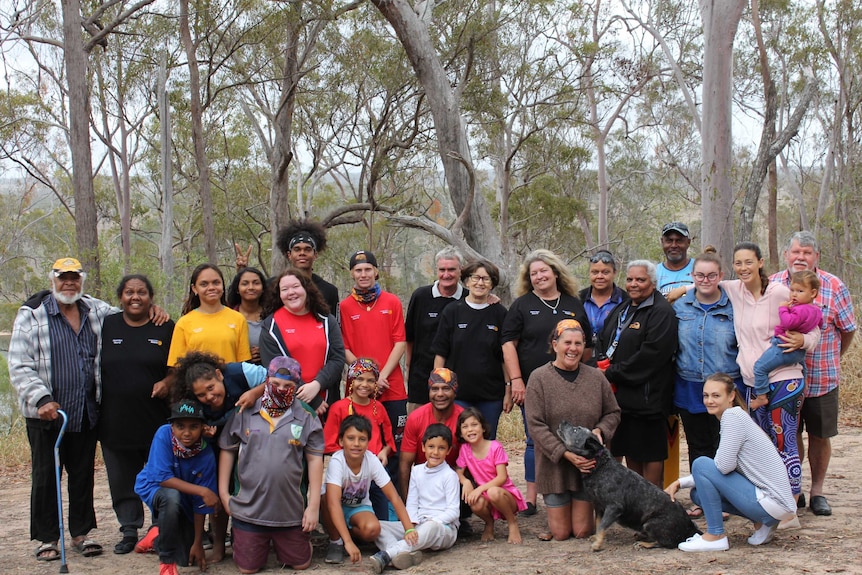 A group of people, both Indigenous and non-Indigenous, and of all ages, stand and kneel, smiling, in a bush setting.