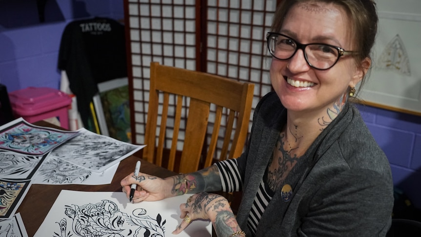 Mim d'Abbs is drawing an image of a woman's face. She is smiling at the camera and has many tattoos.