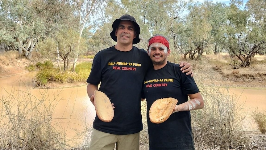 Two men standing by a river holding coolamons