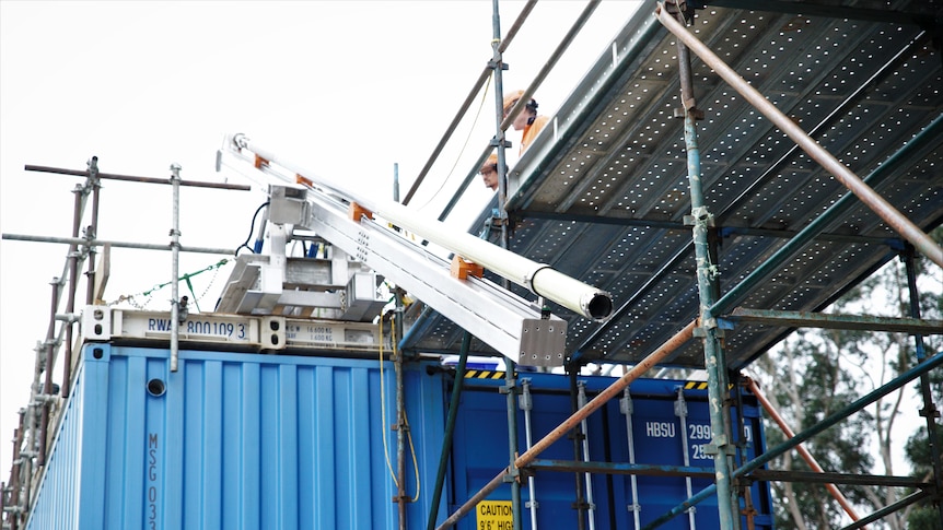 Two people stand on a scaffold above a shipping container and a 10 metre ice drill