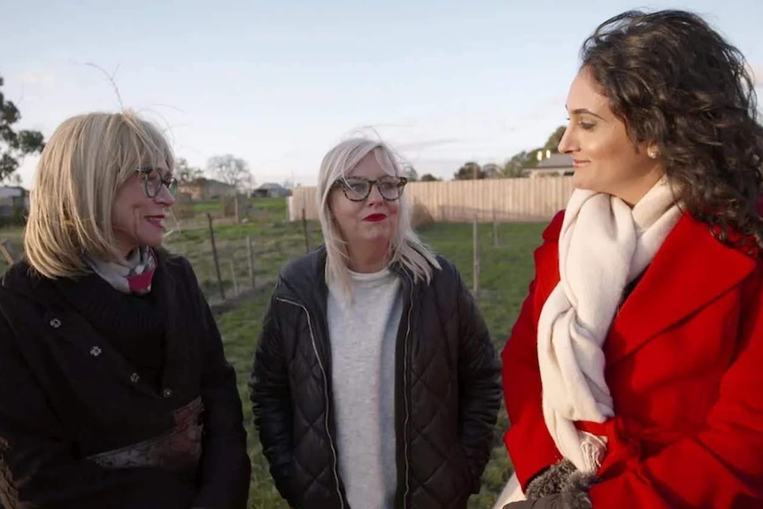 Three women in coats stand in a fenced paddock