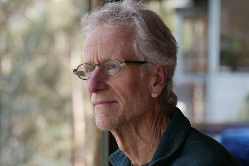A white-haired man wearing glasses leans on a balcony railing surrounded by bushland.