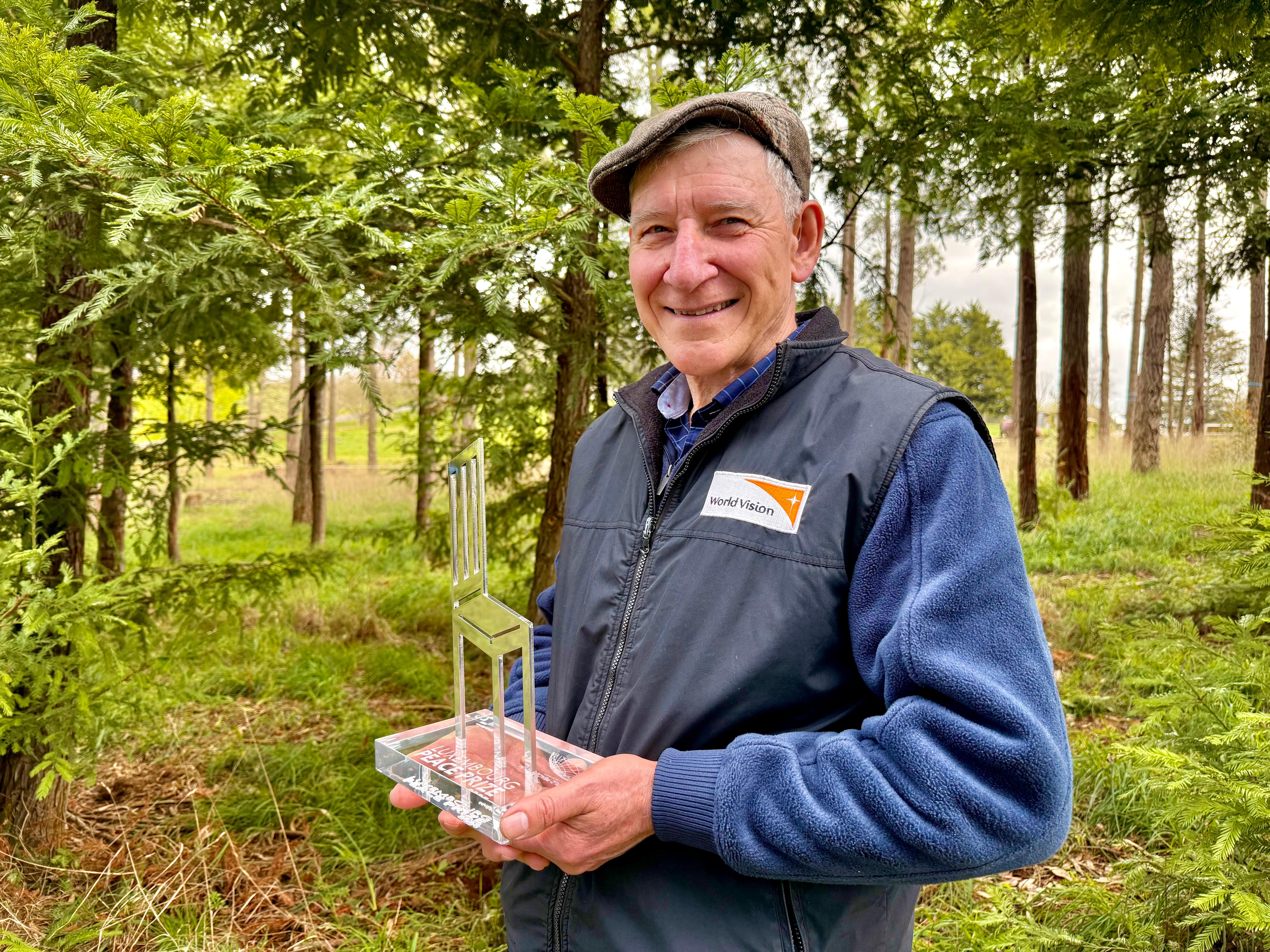 A man standing in a grove of trees, holding a metal award in the shape of a chair in his two hands. 