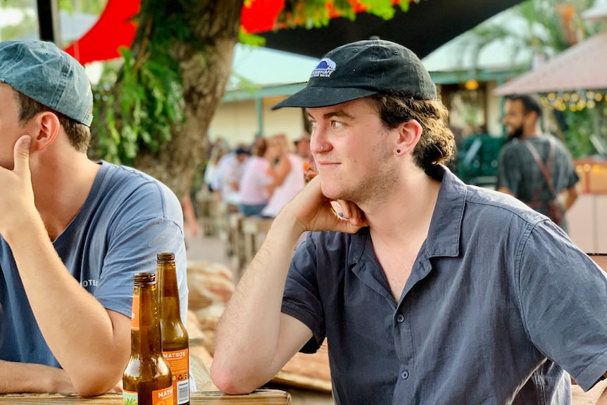 A young man wearing a cap sits at a table with a beer in front of him