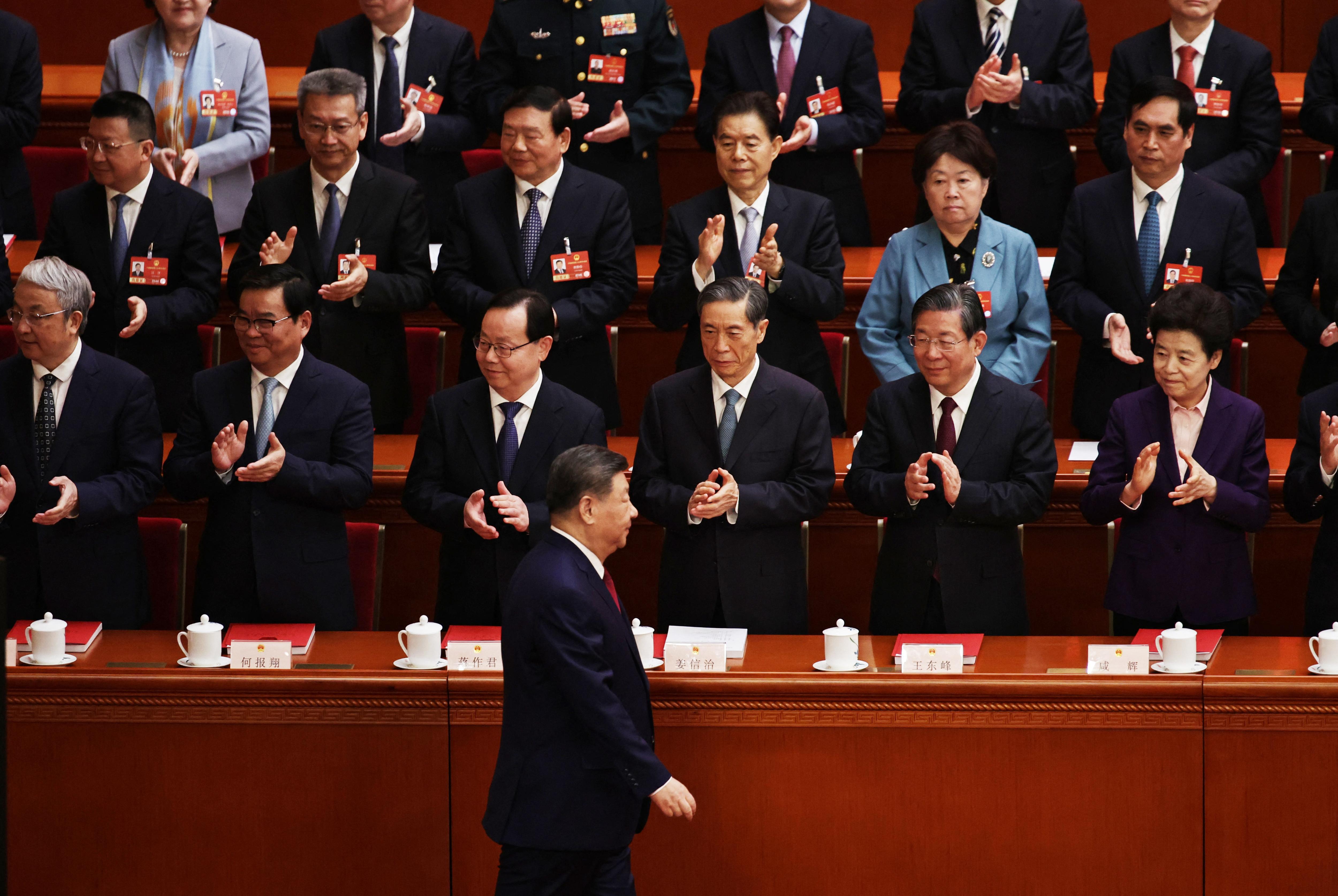 A man in a dark suit and red tie walks past rows of clapping officials.