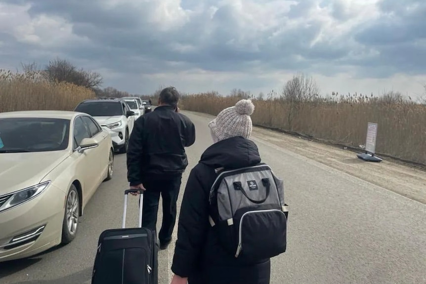 A man and a woman walking along a road with their suitcases while a queue of cars travels in the opposite direction