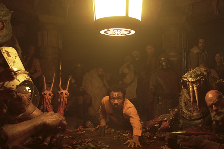 Still from Solo: A Star Wars Story of Donald Glover reaching over a poker table in a dark room surrounded by creatures
