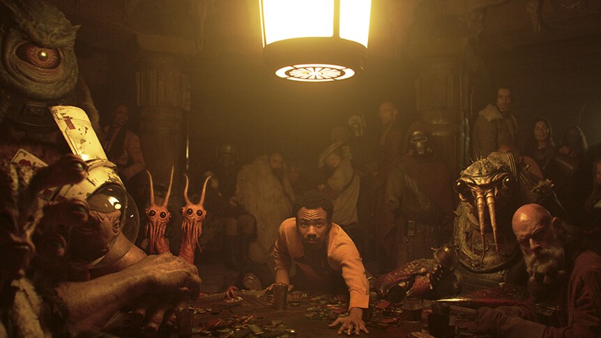 Still from Solo: A Star Wars Story of Donald Glover reaching over a poker table in a dark room surrounded by creatures