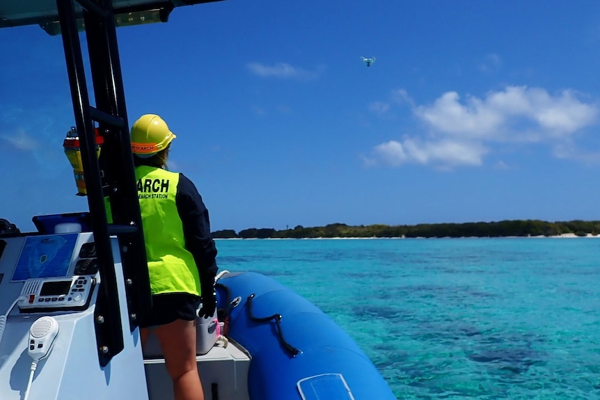 A woman in a yellow vest on a boat in blue waters, looking up at a drone