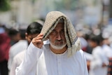 A bearded man in Islamic garb holds a folded decorative cloth on his head, casting a shadow over his face and upper chest