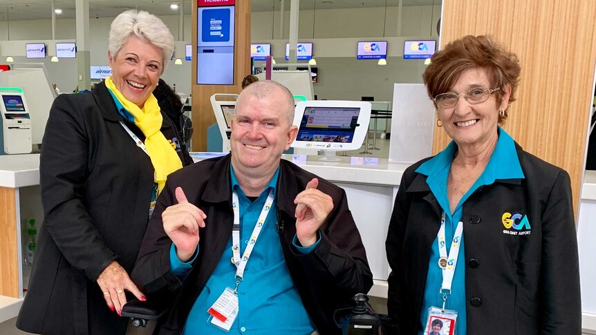 Brett Morris is smiling as he sits between two Gold Coast Airport volunteers behind a desk.