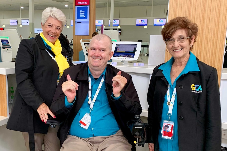 Brett Morris is smiling as he sits between two Gold Coast Airport volunteers behind a desk.