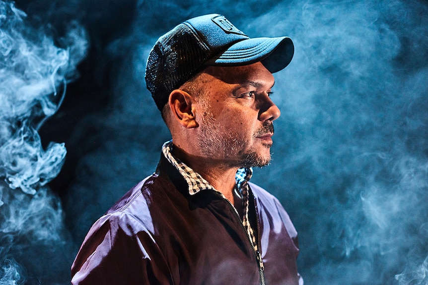 A man wearing cap and jacket looks to the right and stands amongst smoke and dramatic lighting.