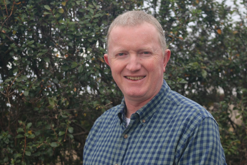 Gene Hodgins - A photo of a middle-aged anglo man in checked shirt smiling, standing in front of a tree