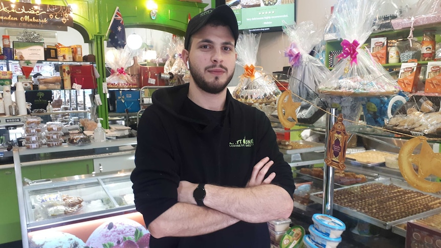 A young man stands in a store with sweets behind him, he is in a black uniform.