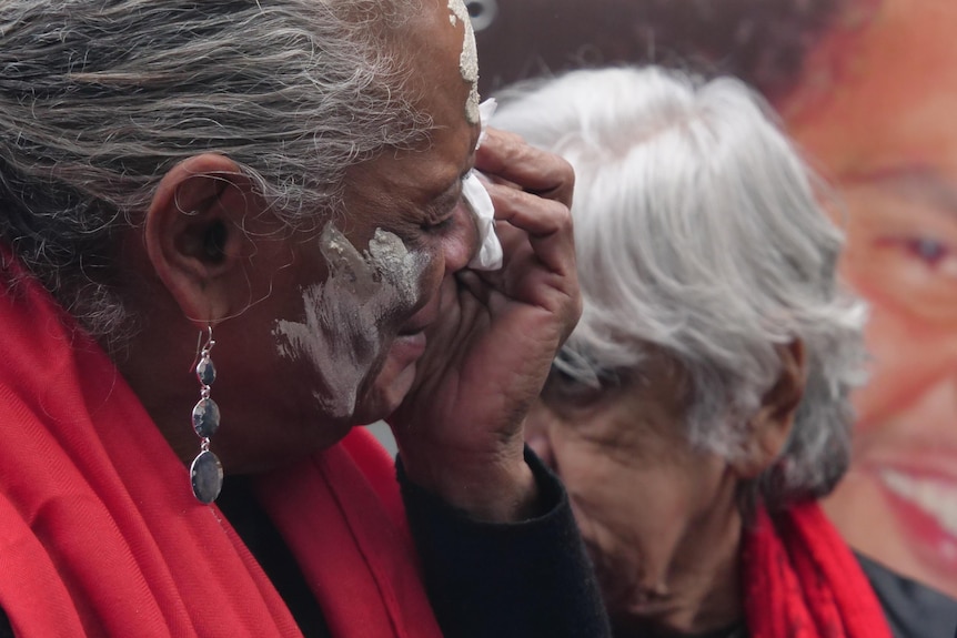 A close up shot of a woman in a red shawl holding a tissue to her eyes as she cries. 