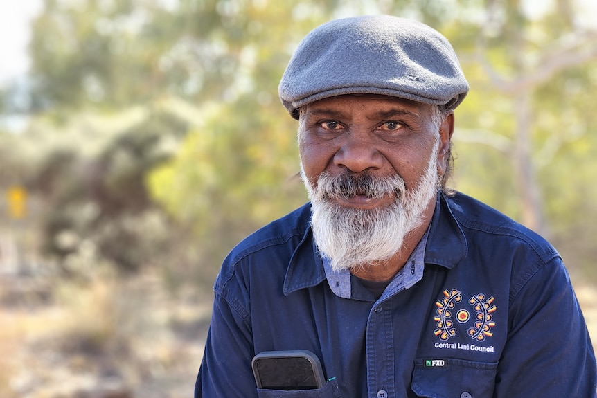 A man in a beret and collared shirt, smiling at the camera, with bushland in the background. 