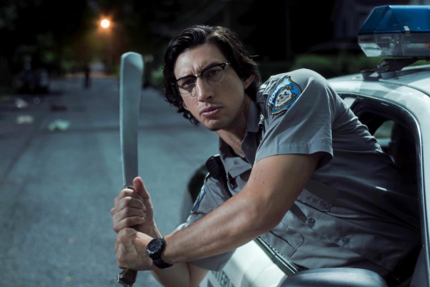 A police officer holds a large knife in a batting position and leans with upper body out of police car window at night time.