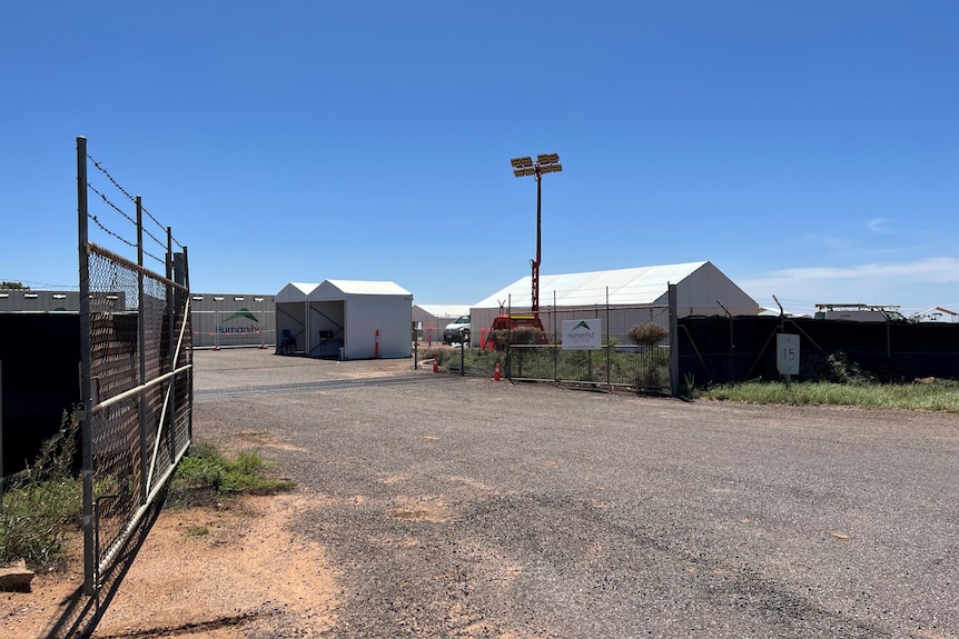 A bitumen driveway leading into a temporary compound of white structures, with an open gate.