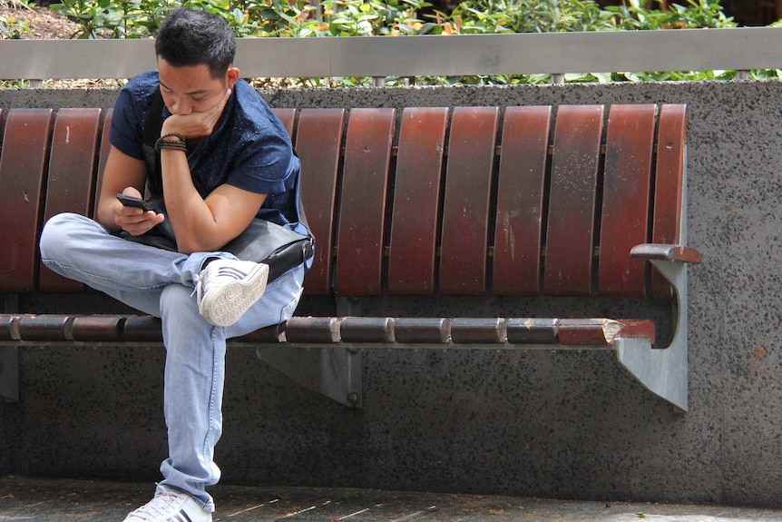 A man reads his mobile phone while sitting on a park bench in Brisbane.