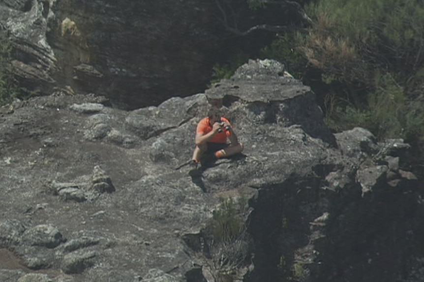 An aerial view of a man sitting on a rock ledge in the Blue Mountains looking through binoculars.