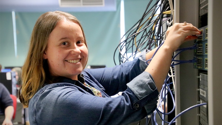 Georgina Barton plugs in cords at a networking rack, smiling.