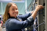 Georgina Barton plugs in cords at a networking rack, smiling.