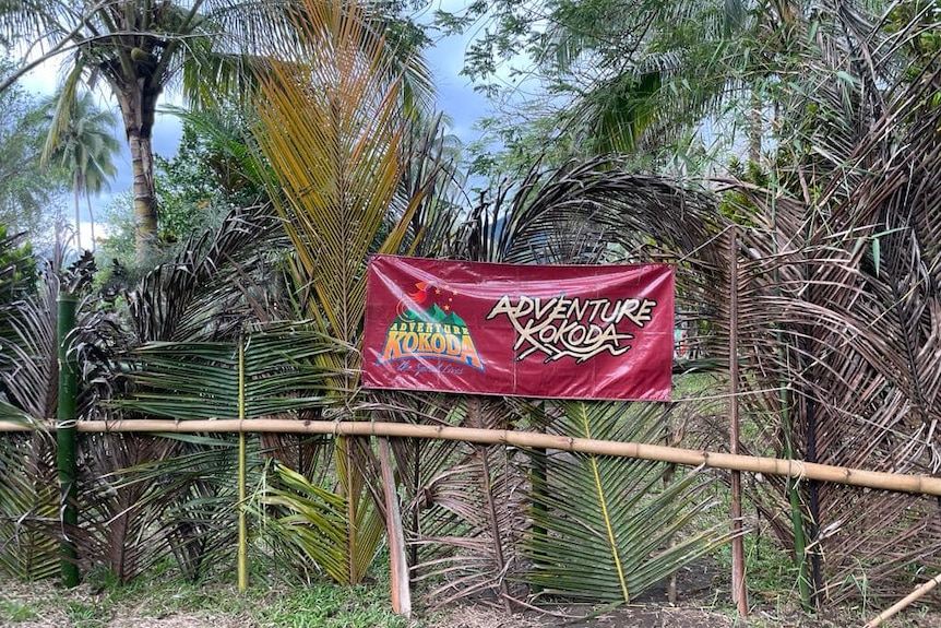 A red sign that says ADVENTURE KOKODA is strung up across palm fronds and branches