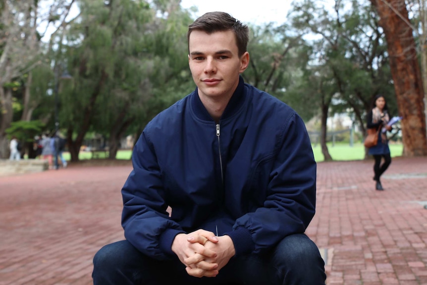 A young man wearing a blue jacket sits outdoors in a brick courtyard in front of trees posing for a photo.