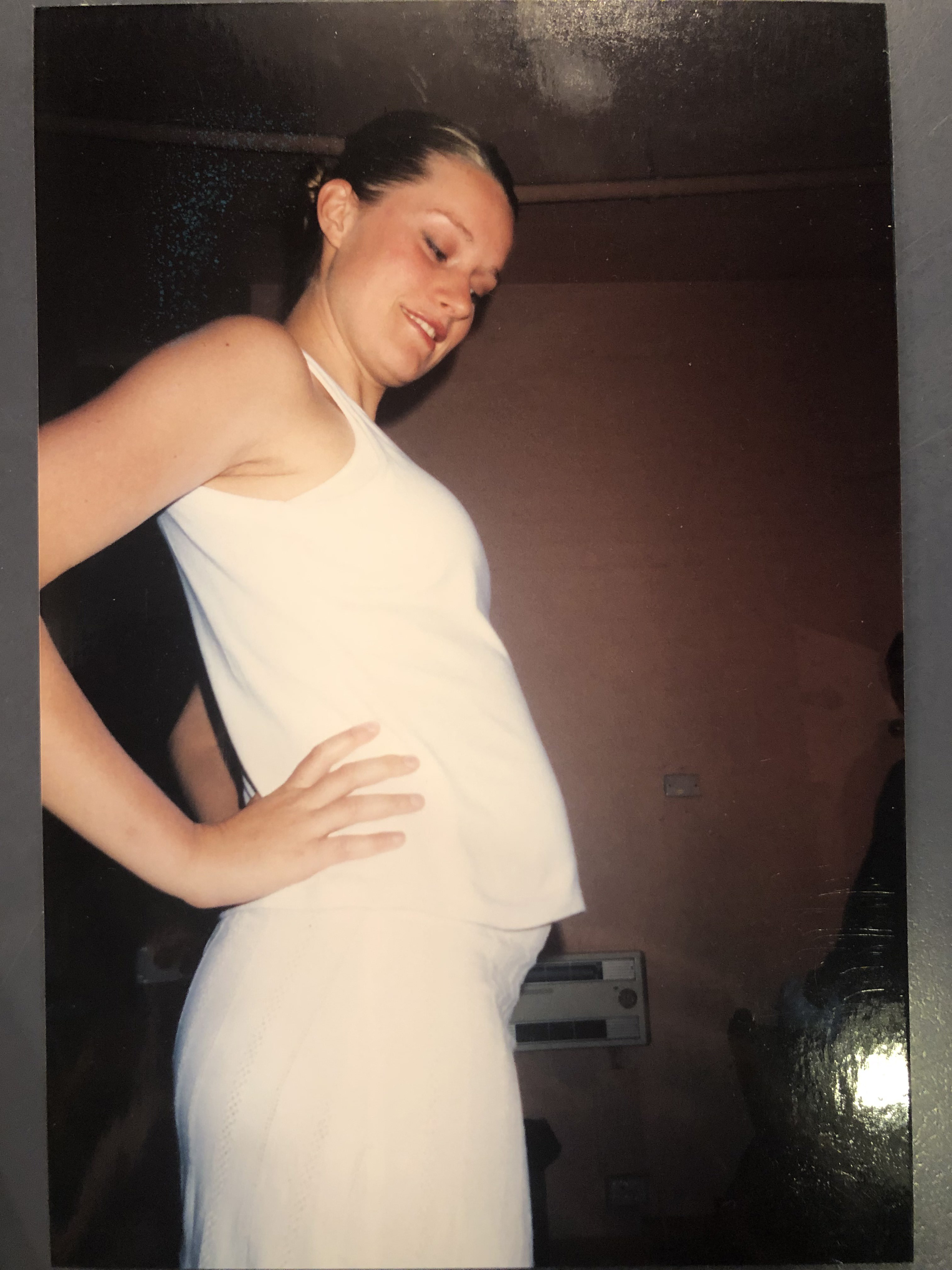 A pregnant blonde white woman with hair pulled into a bun wears a white costume and stands with hands on hips looking at belly.