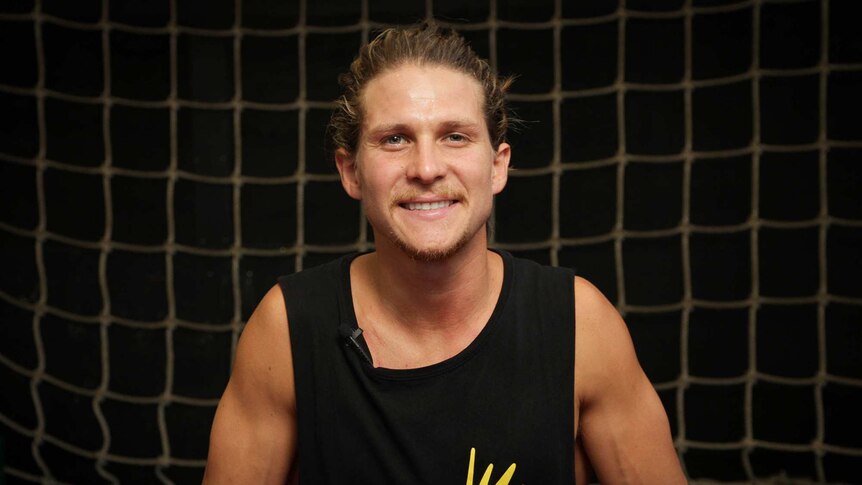 A man with a black singlet in front of a black background with a climbing net.