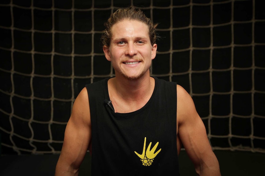 A man with a black singlet in front of a black background with a climbing net.