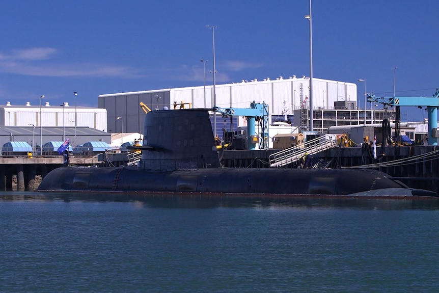A submarine sits docked at shipyards on a fine day. There are cranes and a gang plank over to the sub.
