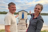 A couple smile as they gesture towards the beautifully restored fishing shed.