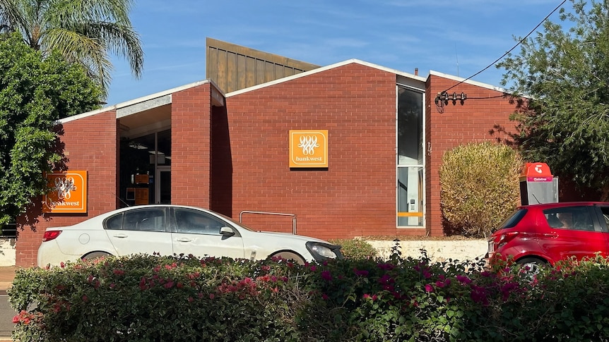 A view across a street of a red brick building with an orange Bankwest logo on the wall. Two cars are parked in foreground. 