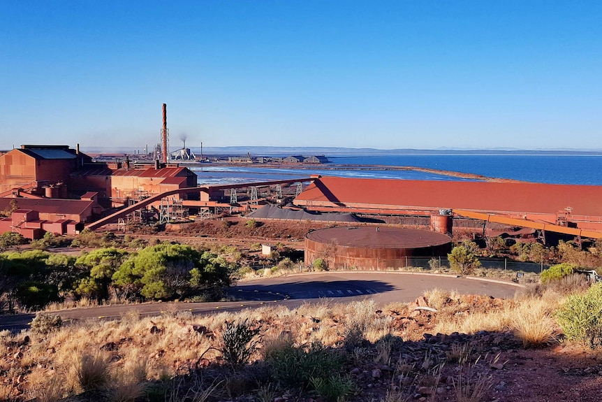 A wide shot of a rusty red coloured group of sheds with dry grass in the foreground and blue sea behind.