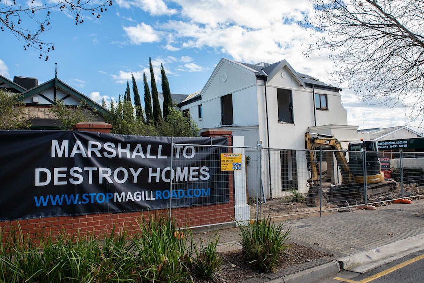 A house under demolition stands next to another house with a sign out front protesting demolitions.