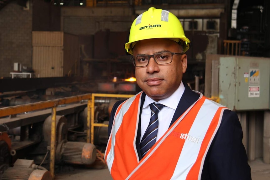 A man in a hard hat and orange vest with a steelworks production line behind him.
