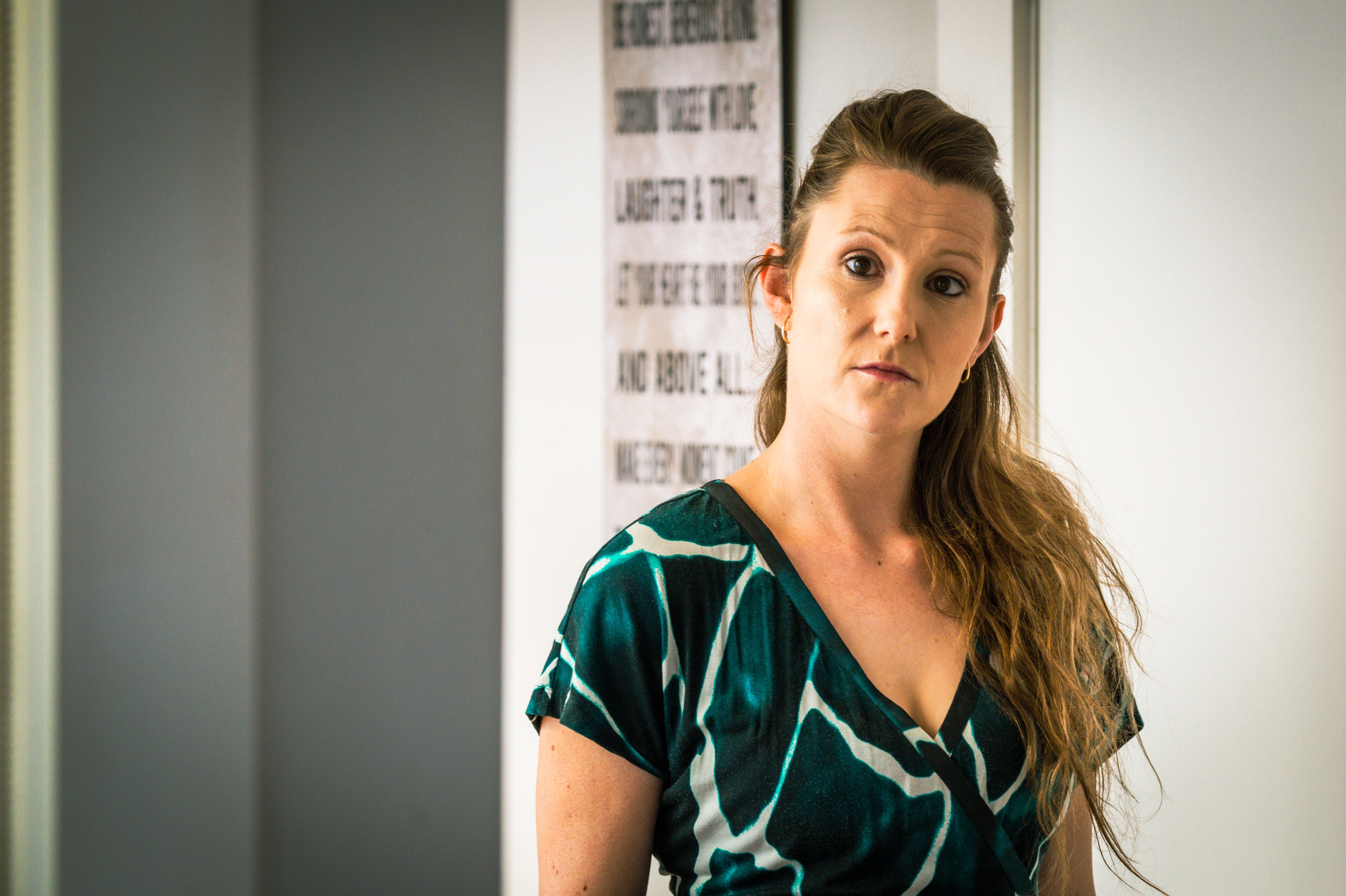 Bree Hughes appears serious, standing inside in a patterned top.