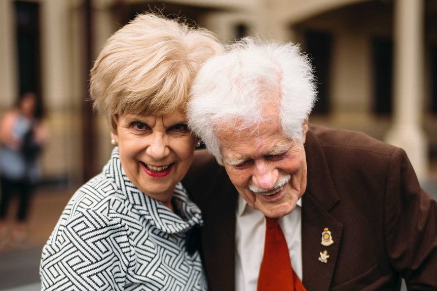An old  man in a brown suit, red tie, and war medals laughs next to a a woman in a white jacket and pearls, also laughing.